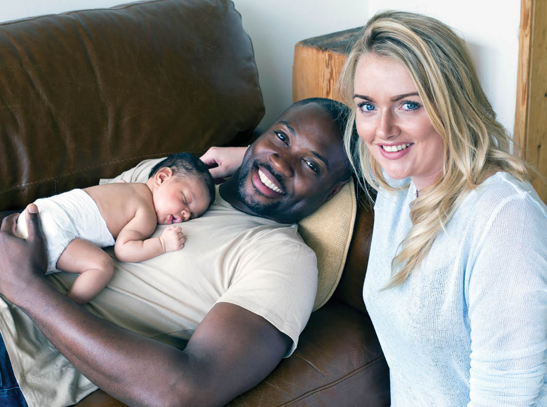A mother and father with their newborn baby daughter at home. They are looking at the camera and smiling.